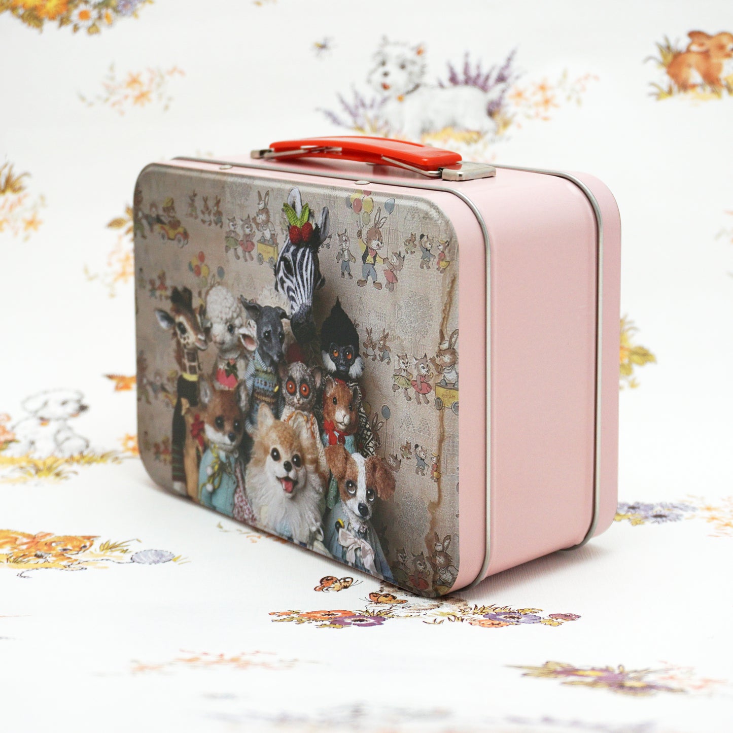 A vintage-style pink metal lunch box featuring a whimsical illustration of various animals dressed in clothes, including a zebra, dog, and lamb. The suitcase is set against a floral-patterned background, adding a playful touch to the scene.