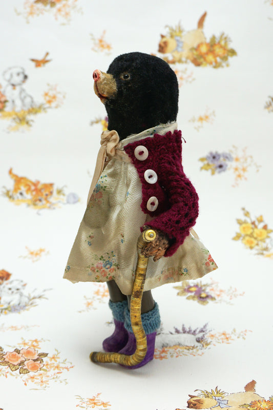 A handmade mole, dressed in a burgundy cardigan, a cream floral dress with a beige ribbon bow, and purple knit socks, holding a curved gold-coloured worm. The toy is posed against a white background with a vintage-style floral and animal print.