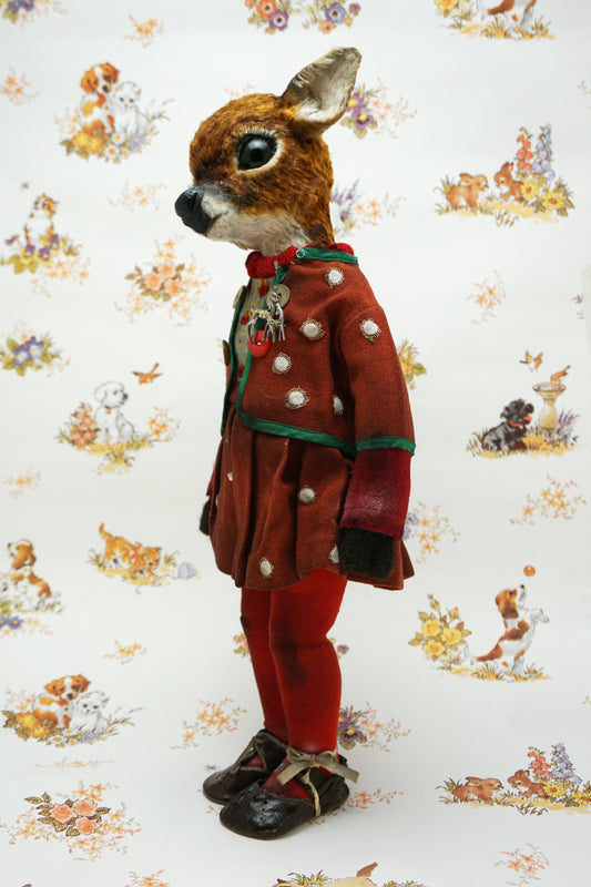 A handmade animal doll with a brown deer head and large dark eyes, dressed in a rust-red velvet jacket with green trim, a knit sweater, red tights, and dark shoes, standing against a white patterned background.