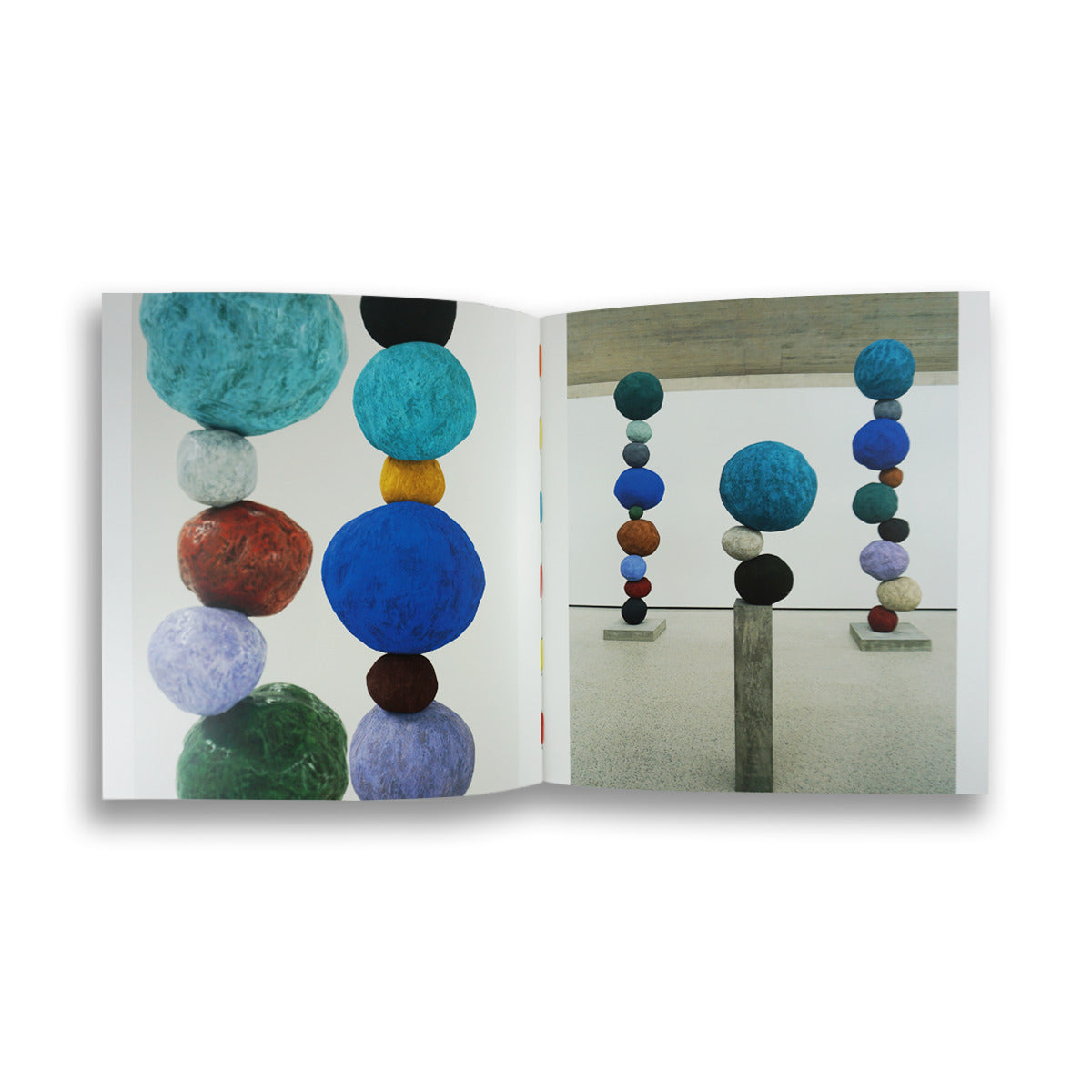 An open book displays two pages featuring vibrant sculptures made of large, colourful spheres stacked vertically. The background shows a minimalist gallery space with a concrete wall and polished floor, enhancing the artwork's visual impact.