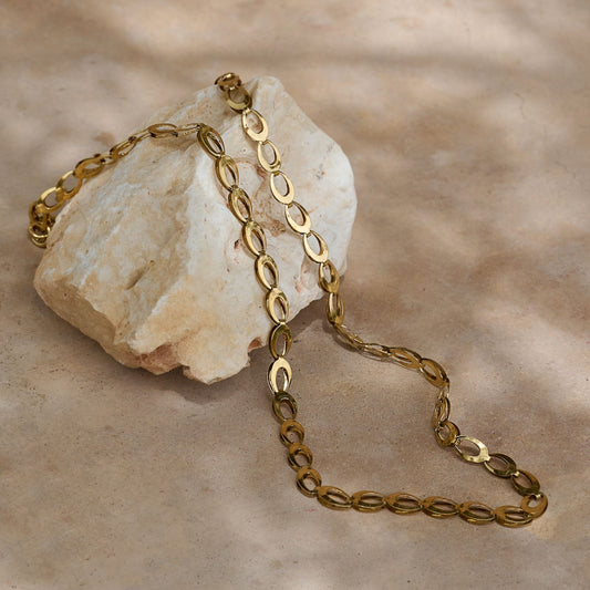 A gold-coloured chain necklace laid out on a textured surface with a stone-like object to the left.