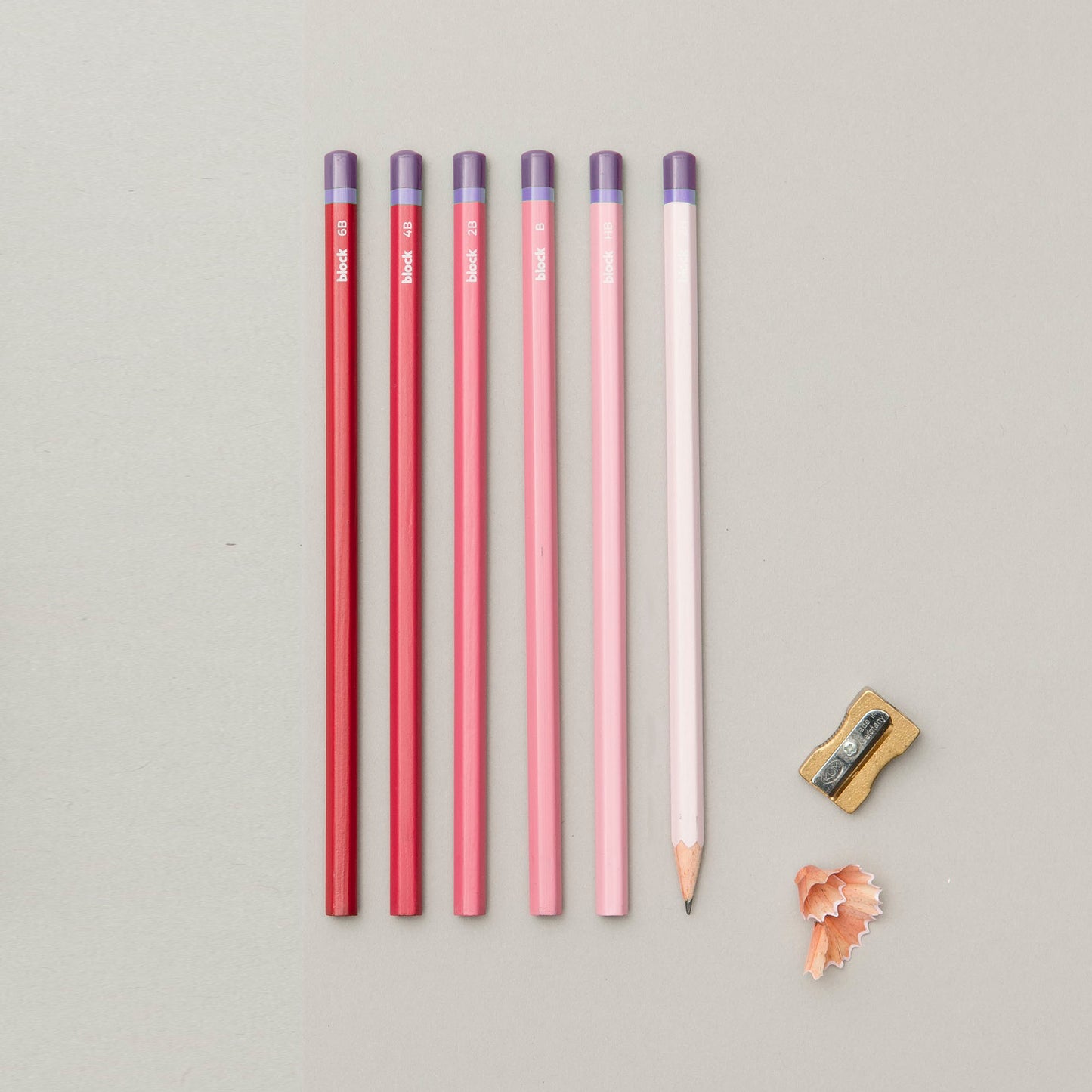 A row of six coloured pencils in shades of red, pink, and white, alongside a small sharpener and pencil shavings on a light green background. The pencils are neatly arranged, showcasing their vibrant colours and clean tips.
