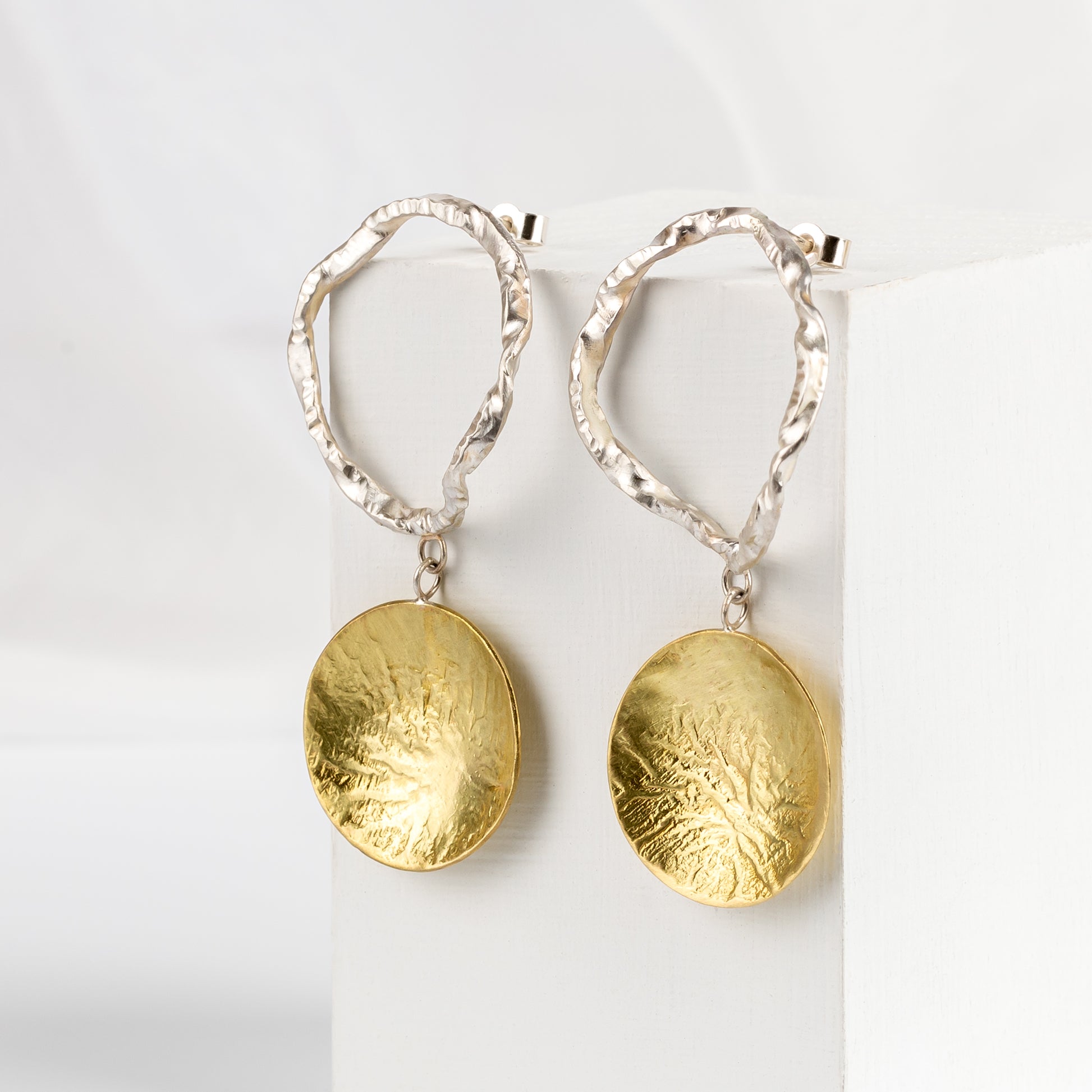 A pair of earrings featuring a silver circular drop and brass disc design, displayed against a white background.