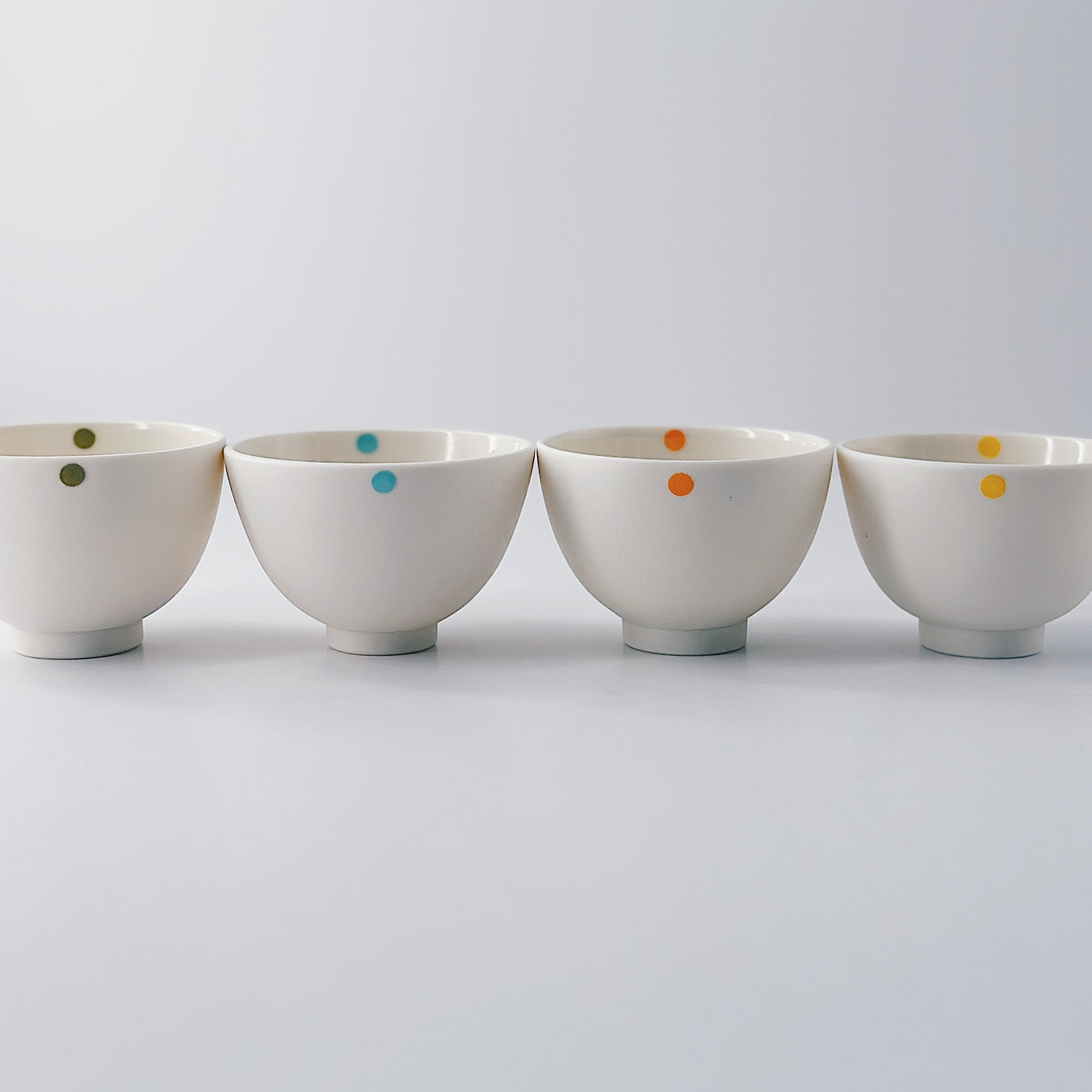 Five small white porcelain bowls each with a single colourful dot arranged in a row on a light grey background