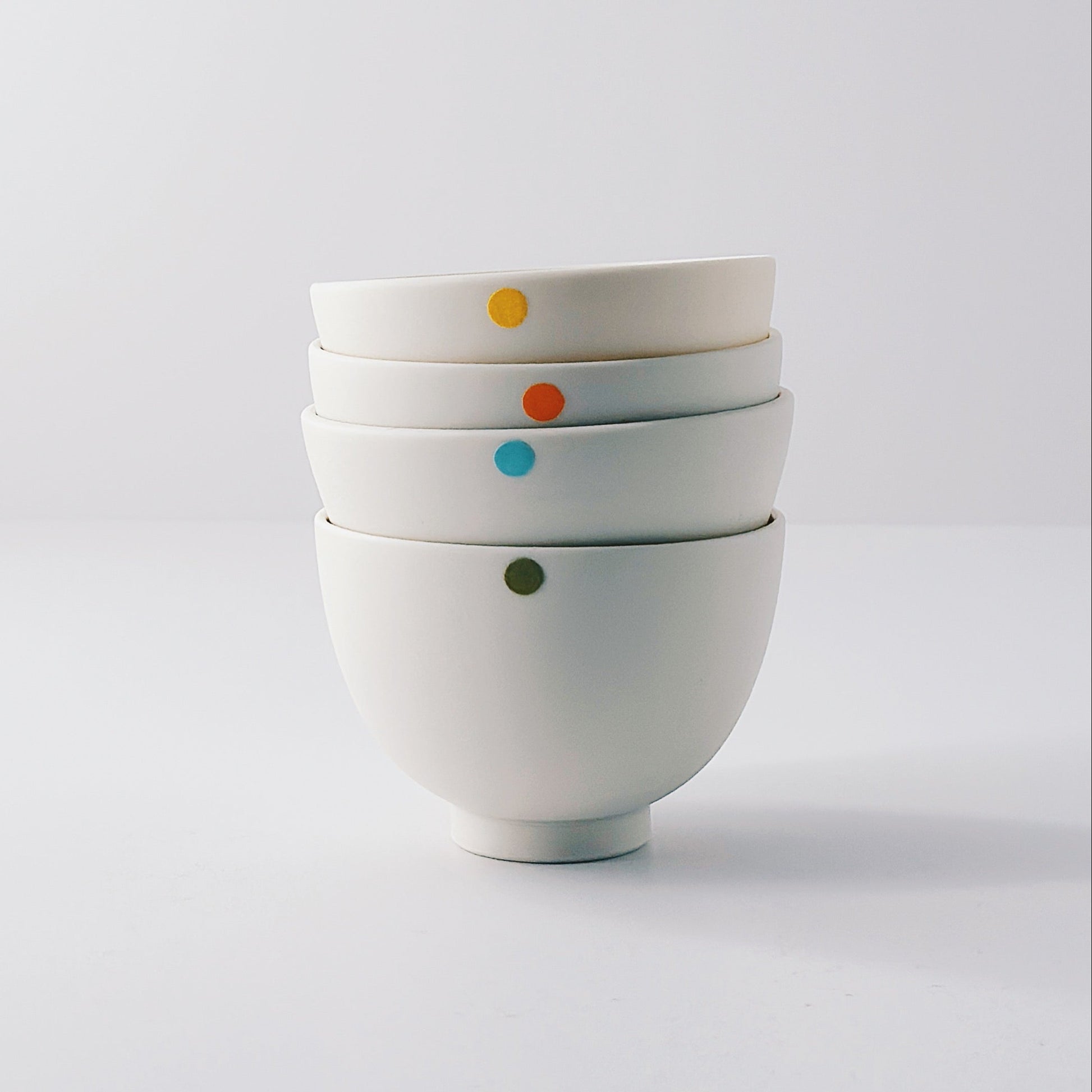 Five small white porcelain bowls each with a single colourful dot arranged in a stack on a light grey background
