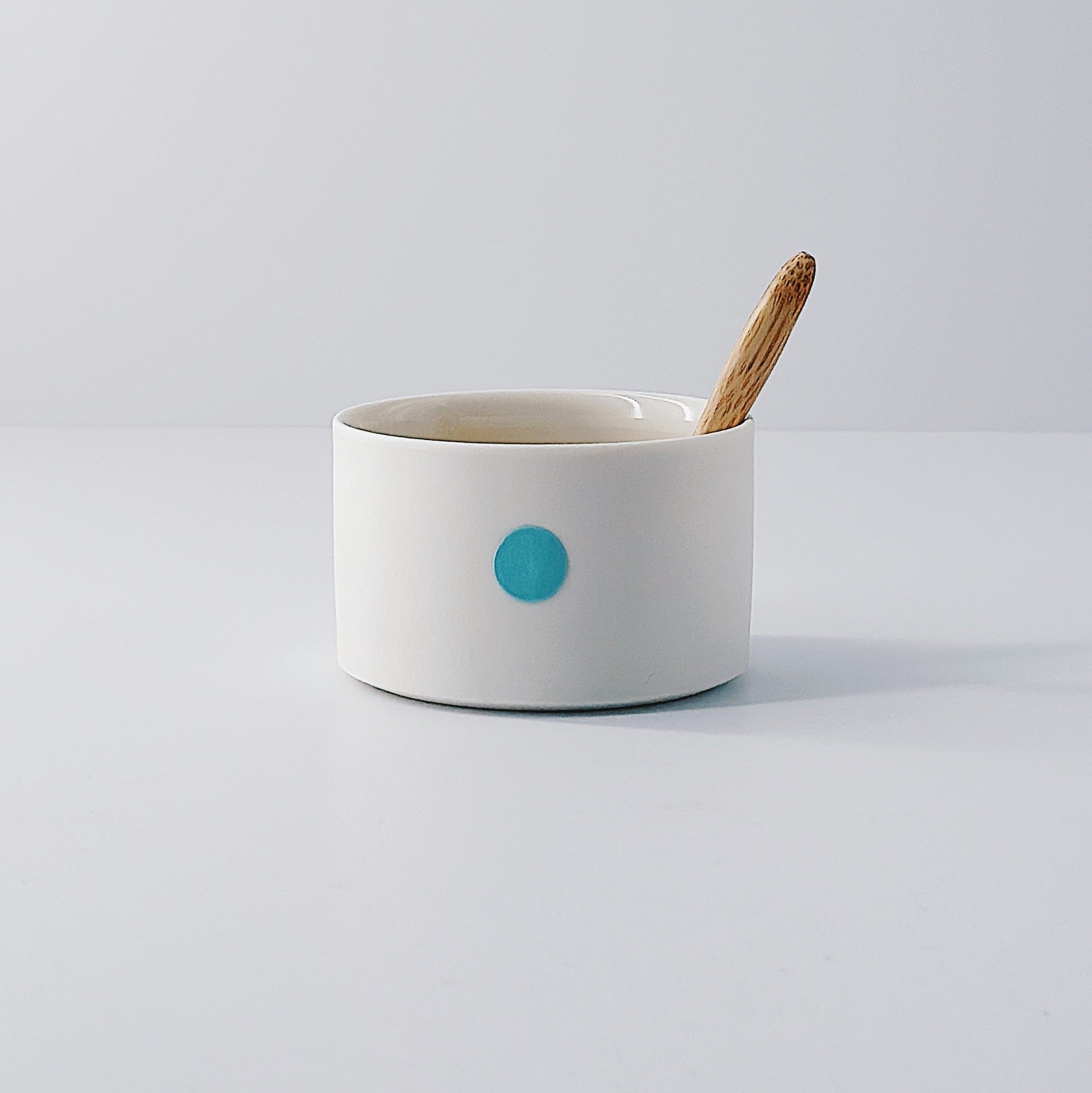 White porcelain pot with a single blue dot and wooden spoon sitting inside on a light grey background