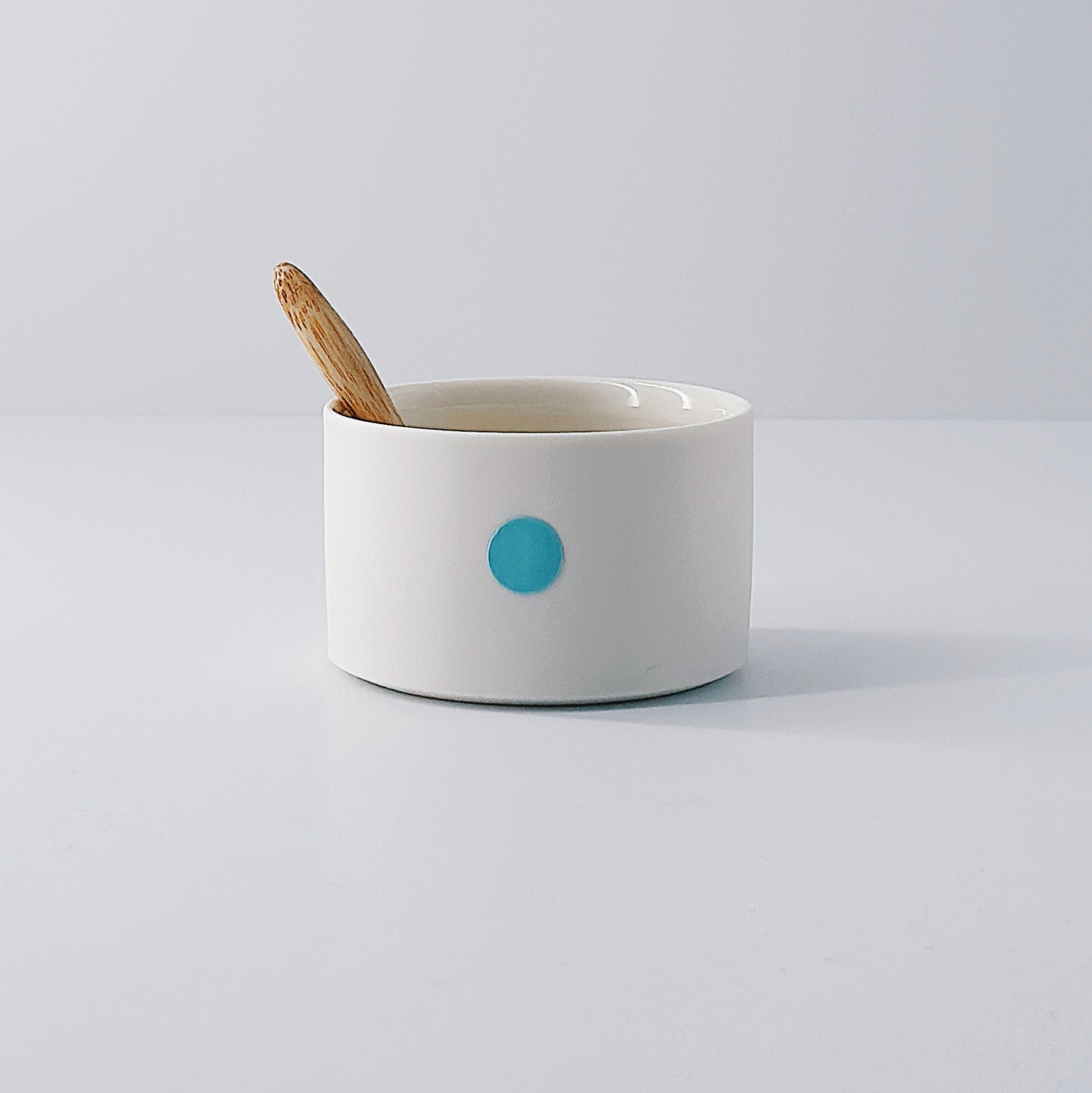 White porcelain pot with a single blue dot and wooden spoon sitting inside on a light grey background