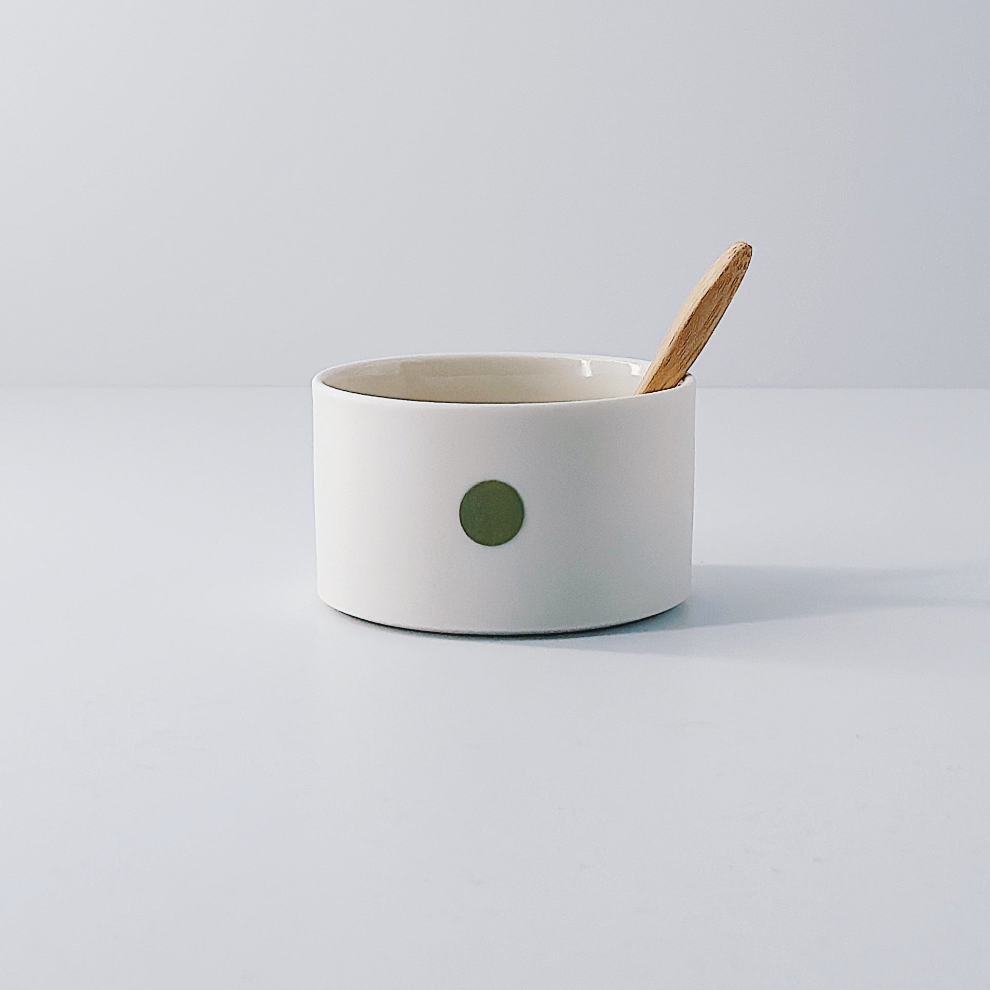 White porcelain pot with a single olive green dot and wooden spoon sitting inside on a light grey background