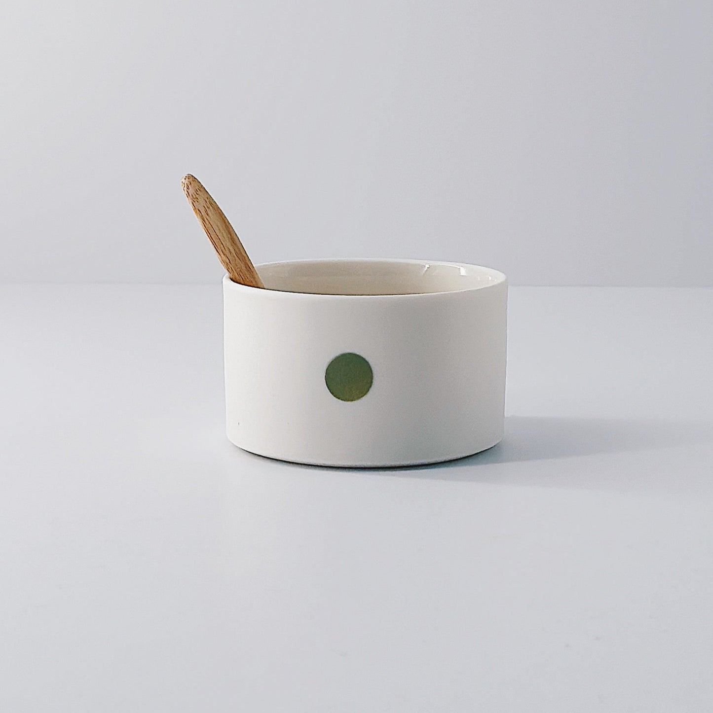 White porcelain pot with a single olive green dot and wooden spoon sitting inside on a light grey background