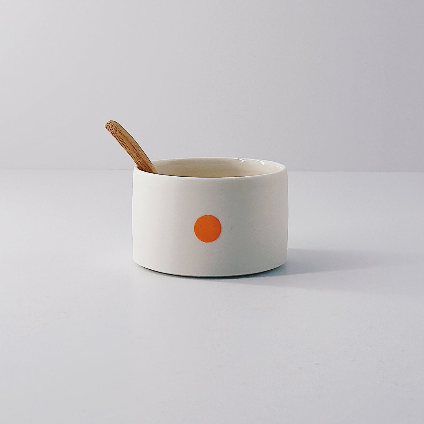 White porcelain pot with a single orange dot and wooden spoon sitting inside on a light grey background