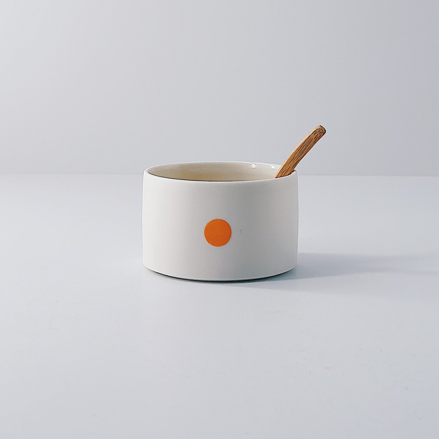 White porcelain pot with a single orange dot and wooden spoon sitting inside on a light grey background