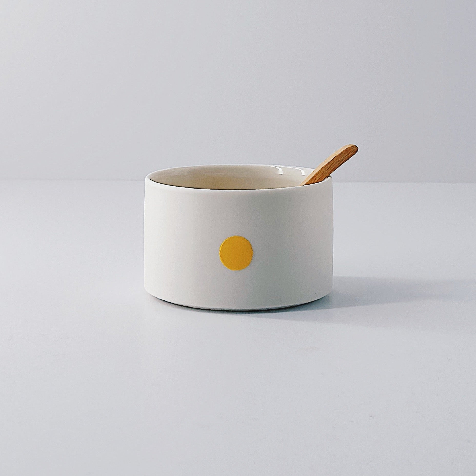 White porcelain pot with a single yellow dot and wooden spoon sitting inside on a light grey background
