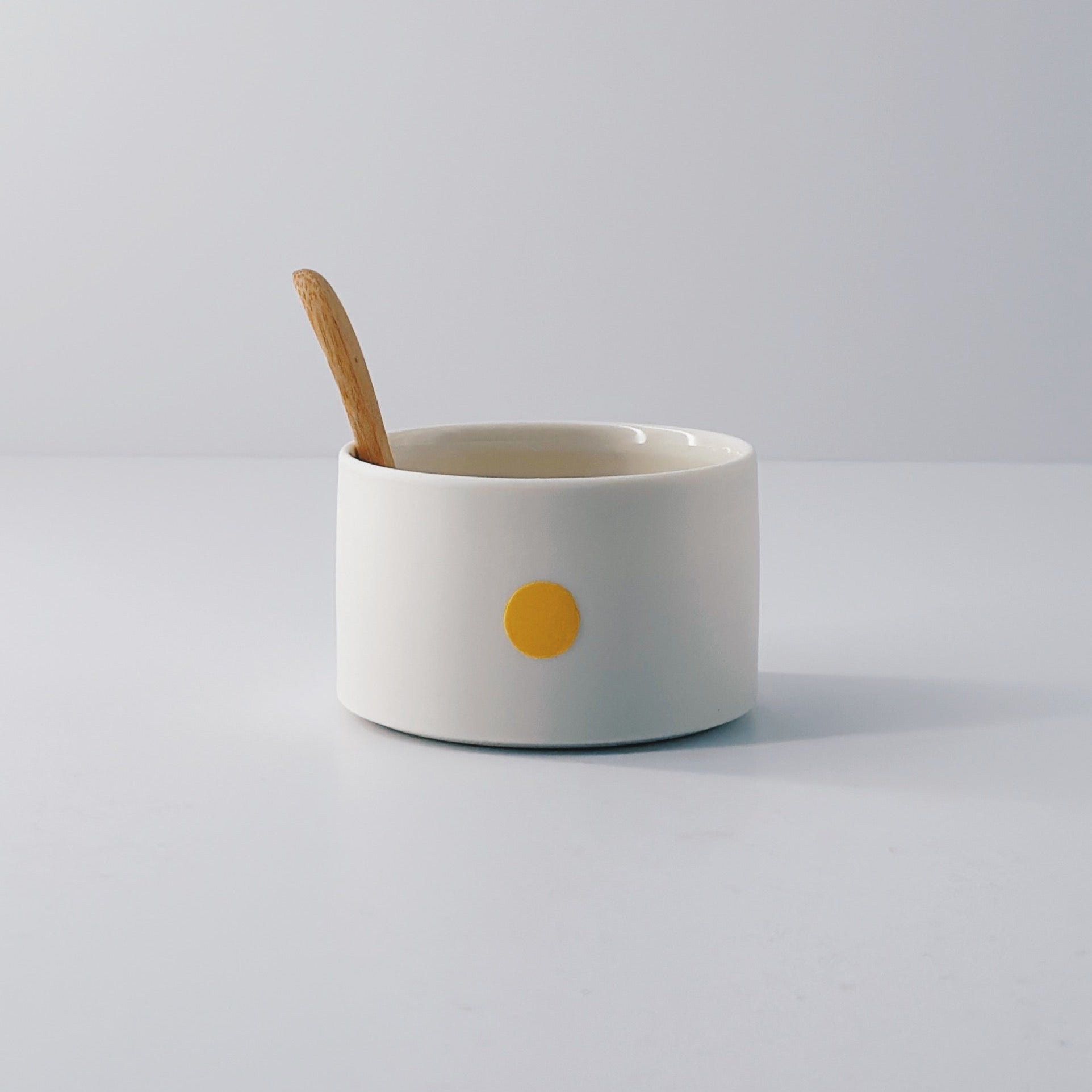 White porcelain pot with a yellow dot and wooden spoon sitting inside on a light grey background