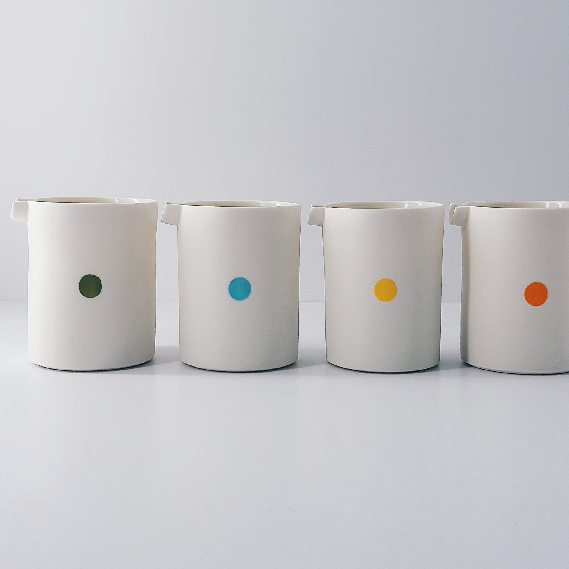 Four white porcelain jugs each with a single coloured dot on a light grey background