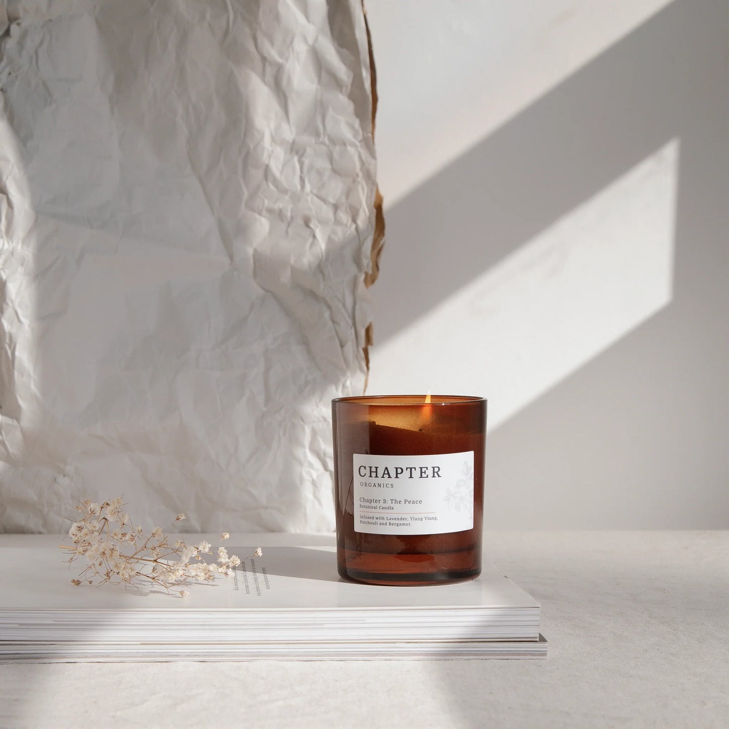 A warm amber candle sits on a stack of white books against a textured, crumpled backdrop. Soft sunlight casts gentle shadows, highlighting the candle's label that reads "CHAPTER" and "Chapter 1: The Peace." A delicate dried flower adds a touch of elegance.