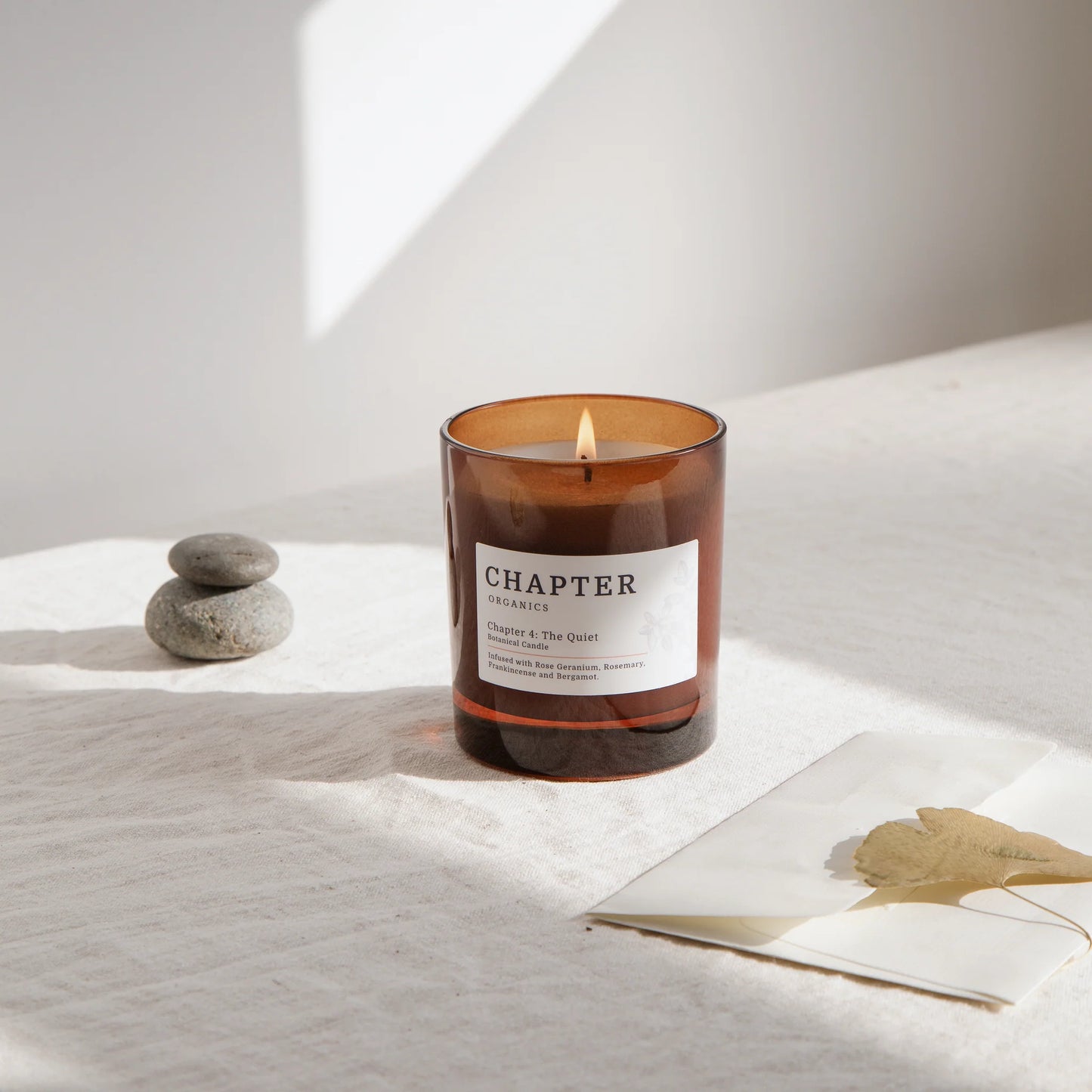 A lit amber glass candle sits on a textured cream surface, accompanied by a stack of smooth stones and a partially visible magazine featuring a leaf. Soft natural light casts a gentle shadow, creating a serene atmosphere.