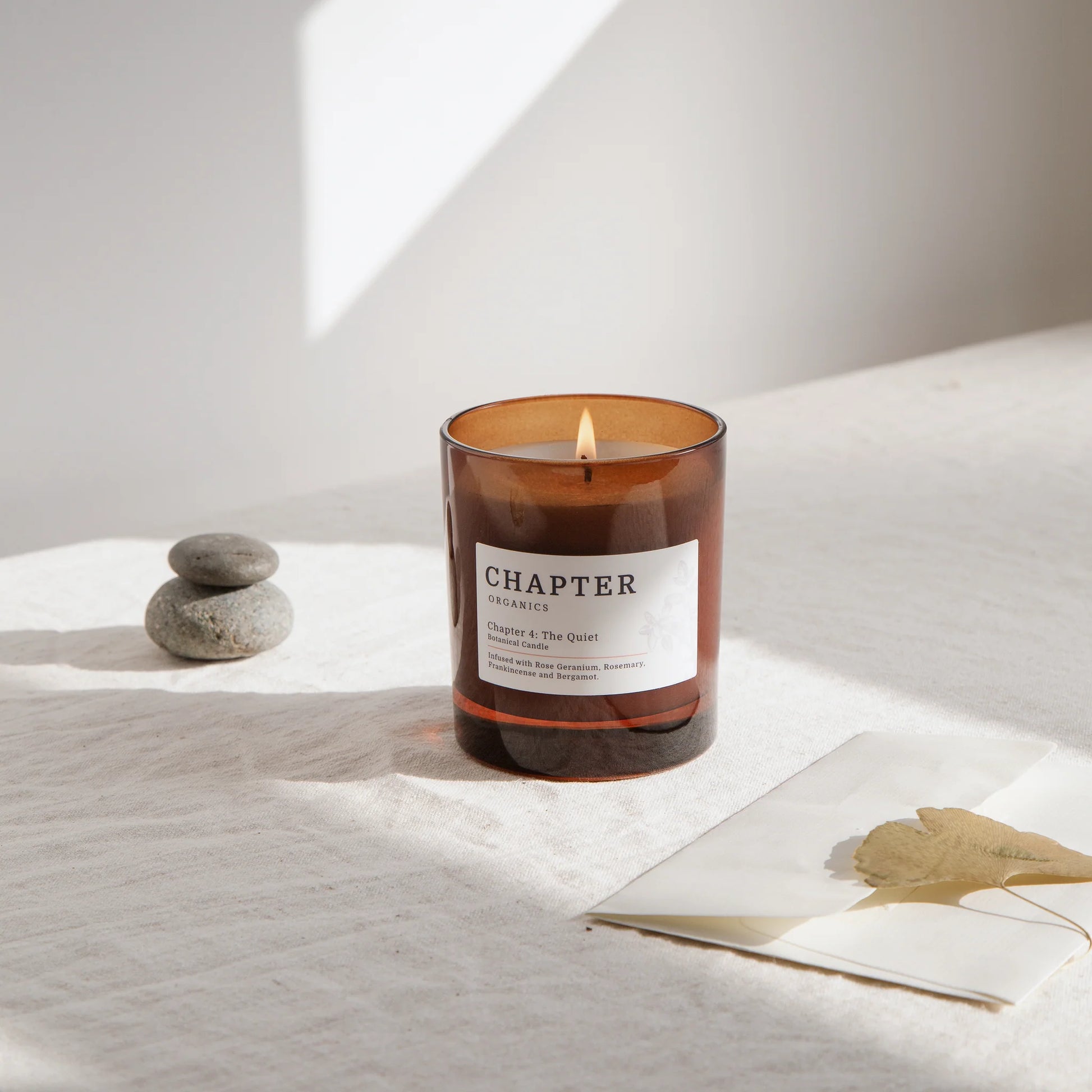 A lit amber glass candle sits on a textured cream surface, accompanied by a stack of smooth stones and a partially visible magazine featuring a leaf. Soft natural light casts a gentle shadow, creating a serene atmosphere.
