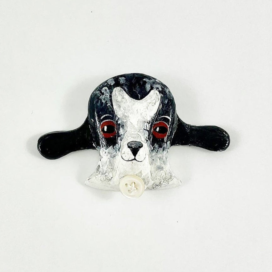 A decorative brooch shaped like a goat’s head, featuring a black and white design. The goat has large red eyes and floppy ears, with a small white button at the neck. The background is plain white, highlighting the brooch's details.