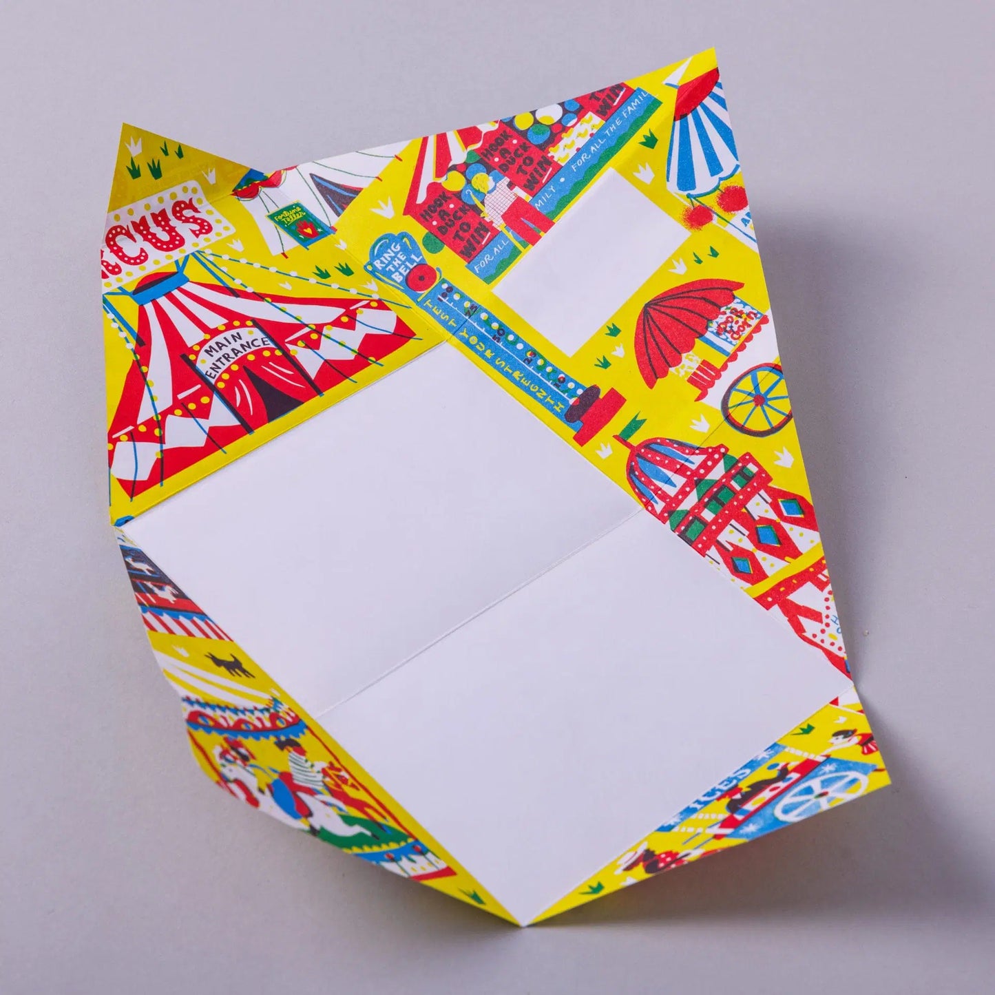 A brightly coloured folded paper featuring a circus theme. The design includes tents, carousels, and various carnival elements on a yellow background. The paper is partially unfolded, revealing a blank white section for writing.