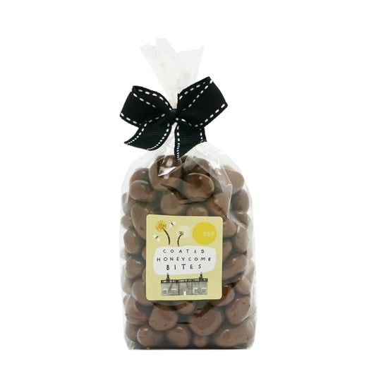 A clear bag of milk chocolate coated honeycomb bites, tied with a black ribbon. The label is yellow and features an illustration of a building and some flowers.