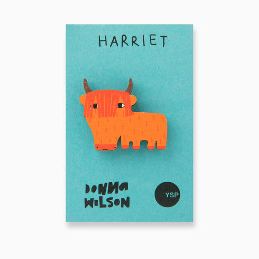 A stylised wooden cut out pin badge in the shape of a highland cow on a blue backing card that features the YSP and Donna Wilson logos.