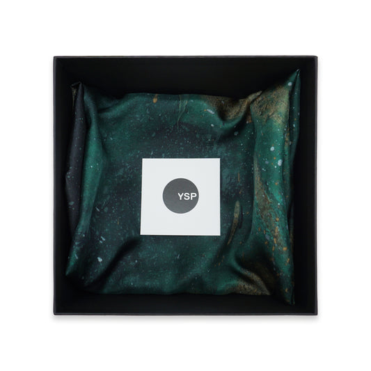 A square black gift box containing a folded dark green and black patterned fabric item with a white square label in the centre that reads 'YSP'.