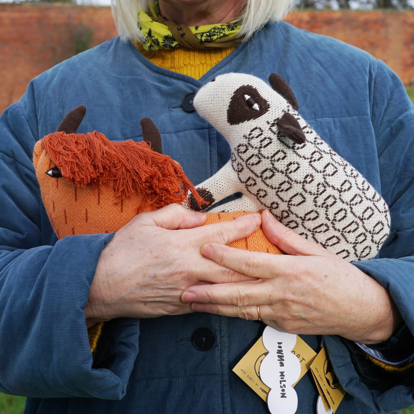 A women standing outside in a blue coat holding a Donna Wilson Wee One sheep and highland cow in her arms.