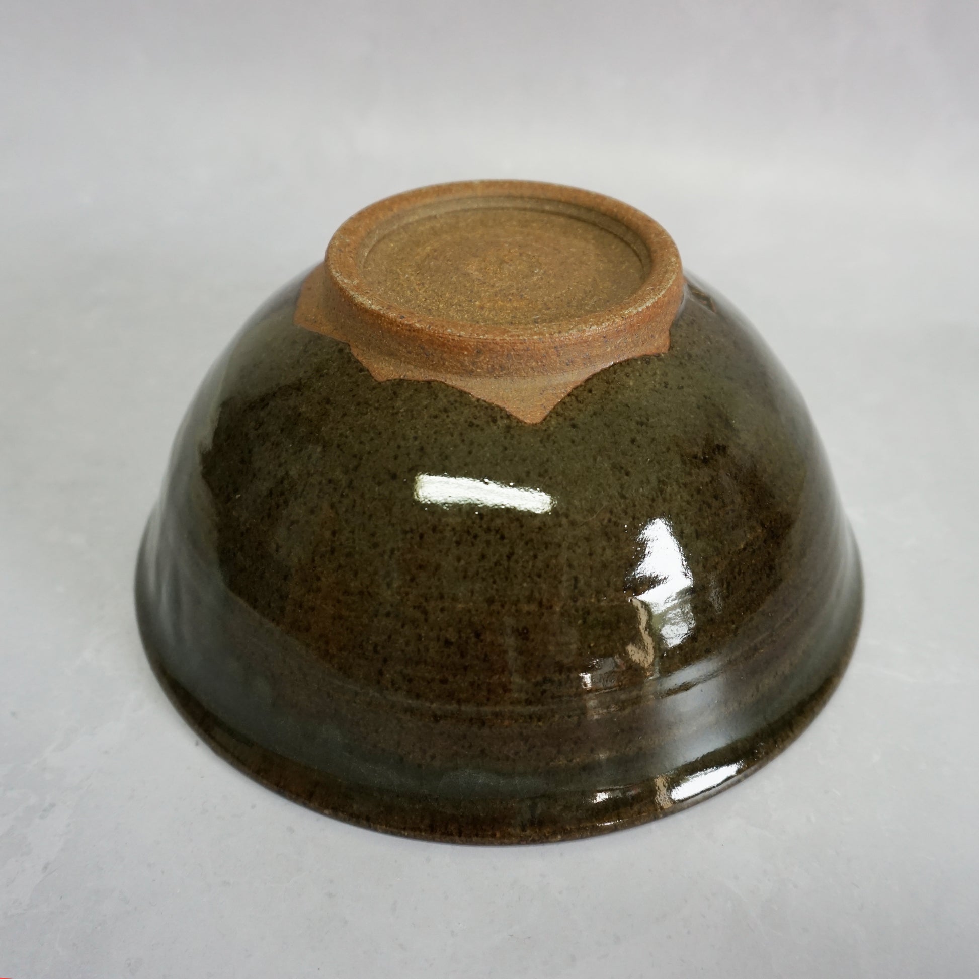 A bottom view of a large stoneware ceramic ramen bowl with a glaze that has shades of grey and green. The glaze is smooth and shiny and the bottom foot of the bowl is brown and unglazed in a circle.