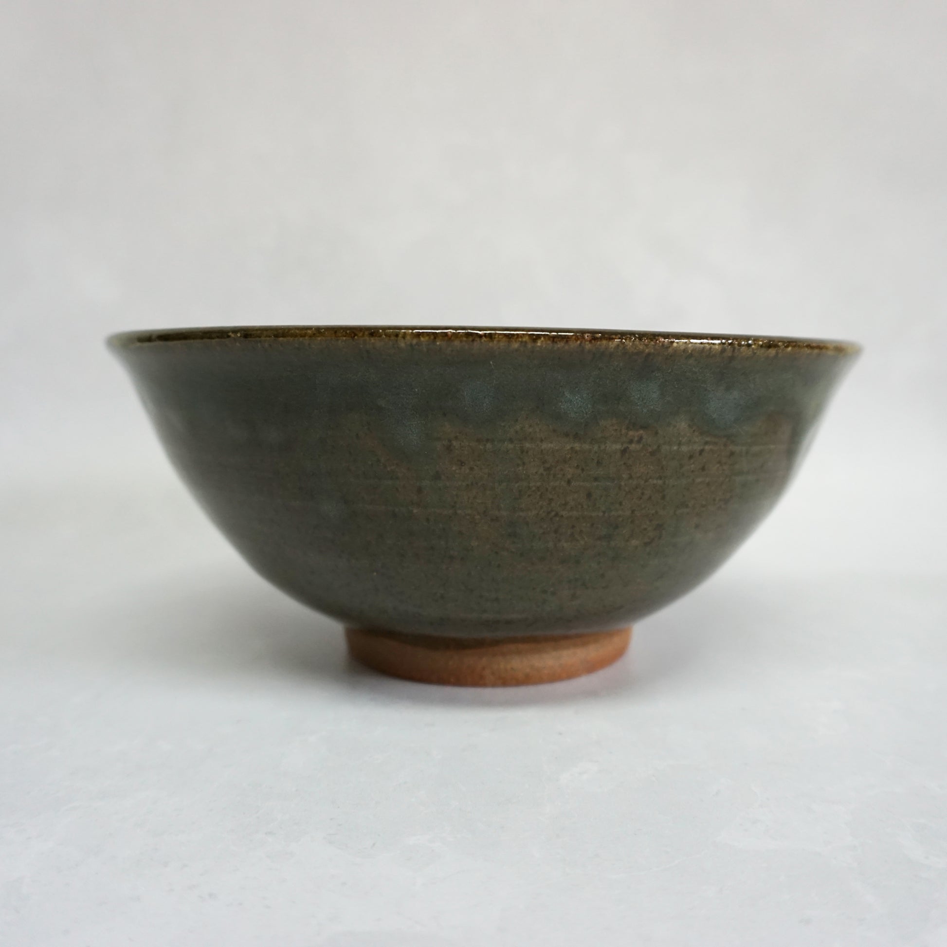 A side view of a large stoneware ceramic ramen bowl with a glaze that has shades of grey and green. The glaze is smooth and shiny and the bowl features a small unglazed foot.