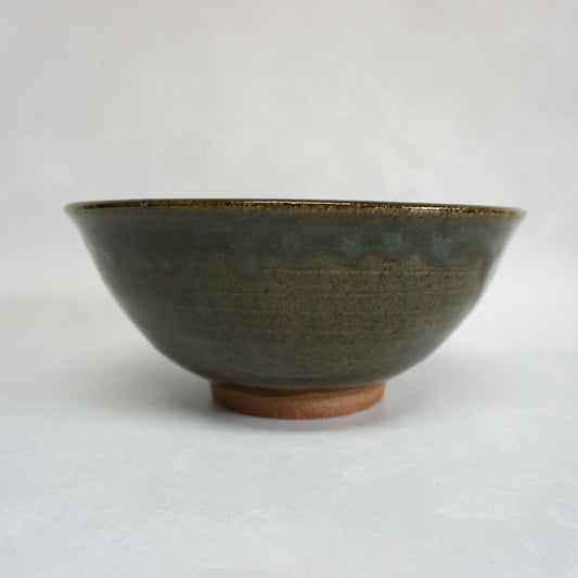 A side view of a large stoneware ceramic ramen bowl with a glaze that has shades of grey and green. The glaze is smooth and shiny and the bowl features a small unglazed foot.