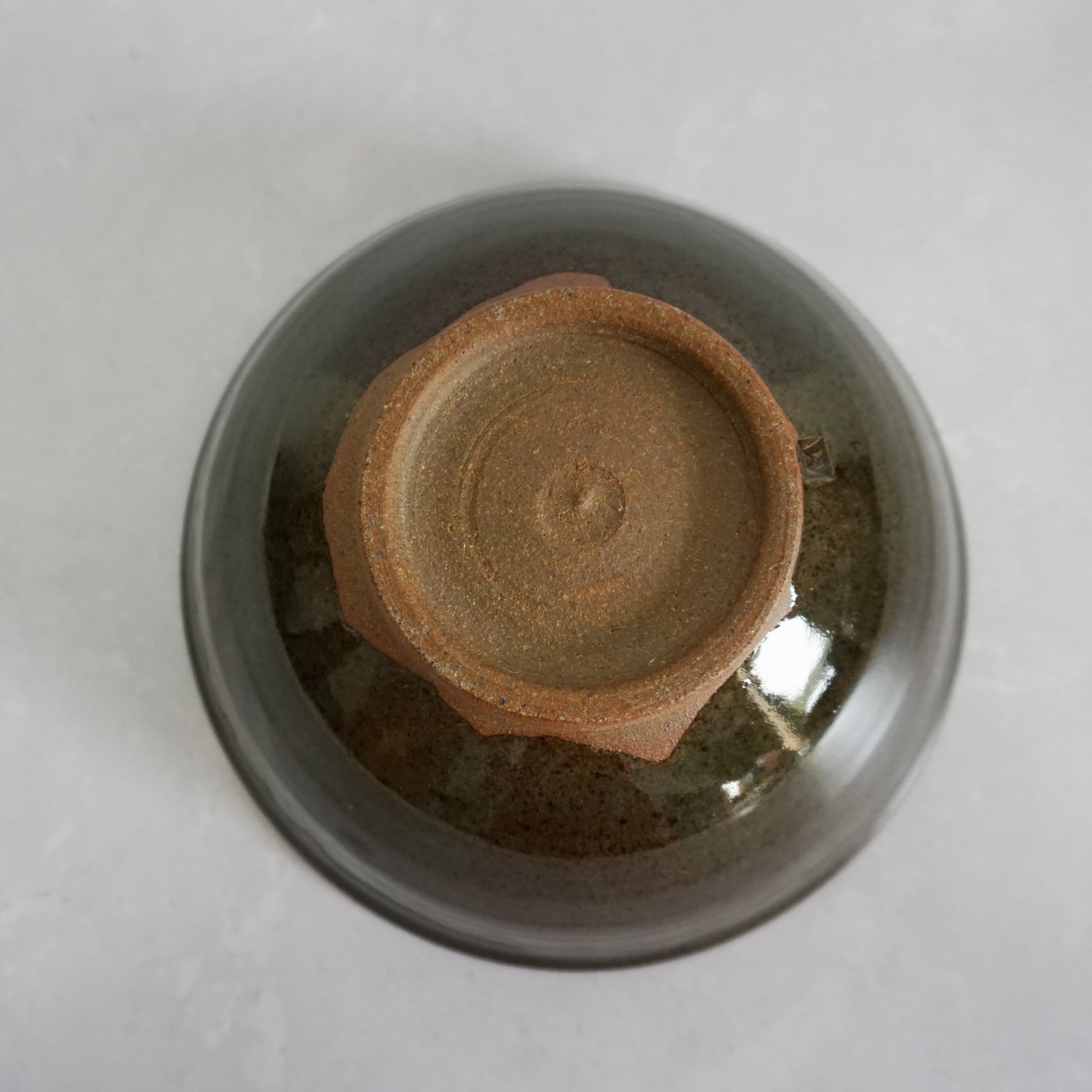 A bottom view of a large stoneware ceramic ramen bowl with a glaze that has shades of grey and green. The glaze is smooth and shiny and the bottom foot of the bowl is brown and unglazed in a circle.