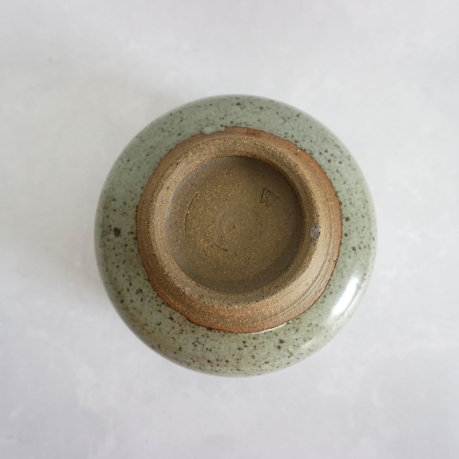 The bottom of a hand-thrown stoneware ceramic tea bowl with a speckled green glaze, featuring the makers stamp.
