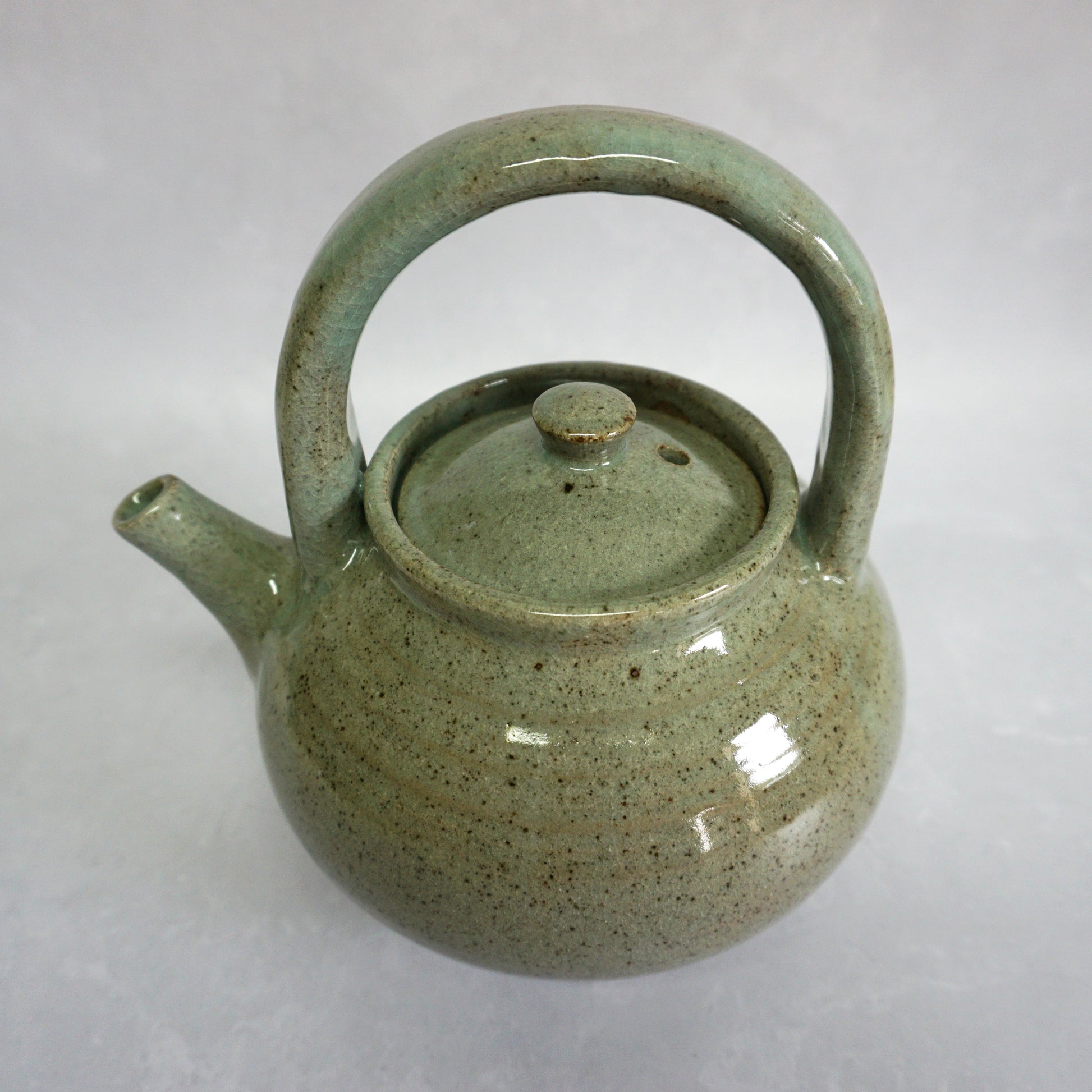 A hand-thrown stoneware ceramic teapot with a green glaze and a curved handle. The glaze is shiny and smooth. The teapot features a delicate spout and lid.