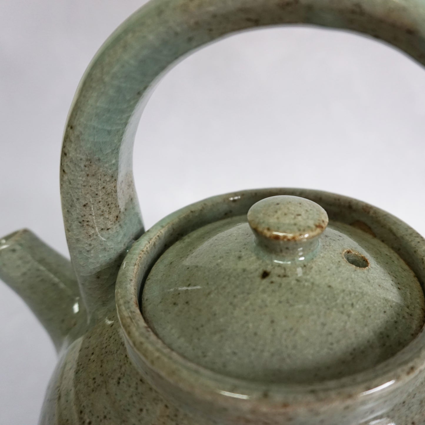 A hand-thrown stoneware ceramic teapot with a green glaze and a curved handle. The glaze is shiny and smooth. The teapot features a delicate spout and lid.