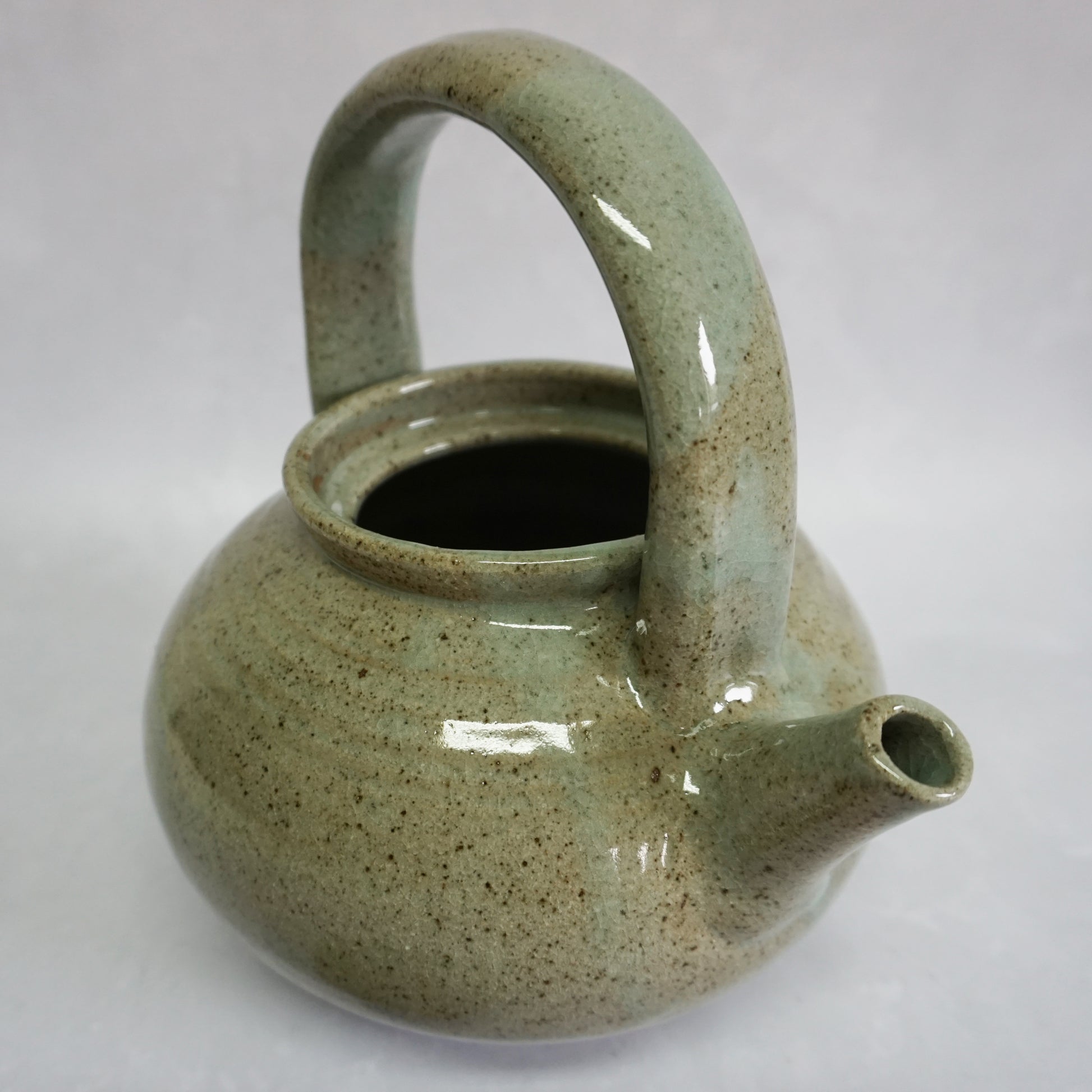 A hand-thrown stoneware ceramic teapot without it's lid. The glaze of the teapot is shiny and smooth. The teapot features a delicate spout.