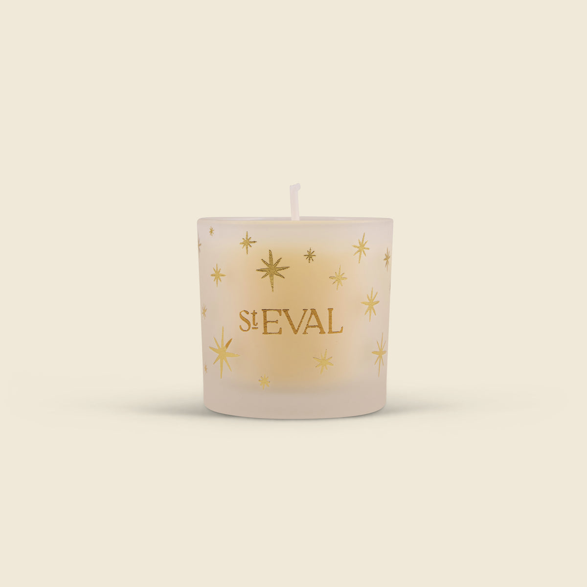 A frosted glass candle with a single wick, decorated with gold starburst patterns and the brand name 'st EVAL' printed in gold on the front, set against a solid cream background.