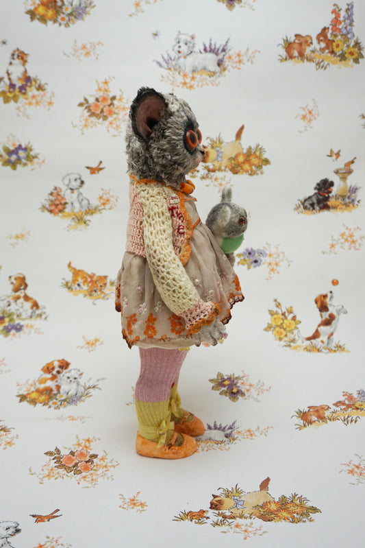 A plush toy resembling a bush baby stands against a patterned background. It wears a knitted dress and holds a small bear toy. The backdrop features whimsical animal illustrations in soft colours.
