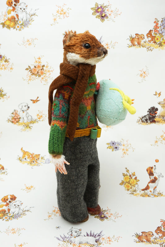 A plush weasel stands upright, wearing a green jumper, brown scarf, and grey trousers. It holds a light blue egg with yellow details. The background features a whimsical pattern of small animals and flowers.