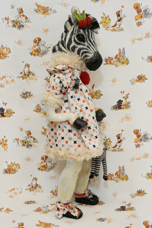 A plush zebra with a patterned dress and red pom-poms stands against a whimsical animal-themed wallpaper. They hold a small zebra toy, and wear black shoes with matching patterned socks.