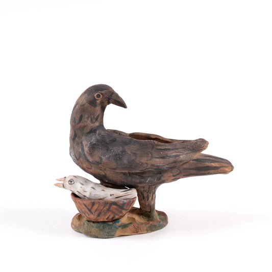 A painted ceramic crow whistle in shades of brown, with a chick in a basket at it's feet. On a plain white background.