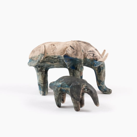 A painted ceramic elephant whistle with a ceramic painted calf. Both are in shades of beige, grey and blue and are standing on a plain white background.