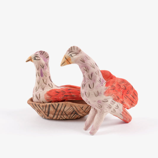 Two painted ceramic birds, one standing and one sitting in a nest, on a white background