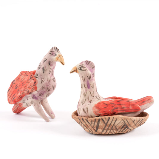 Two painted ceramic birds, one standing and one sitting in a nest, on a white background