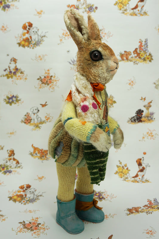 A plush rabbit doll stands upright, wearing a colourful knitted outfit with a rabbit motif. It holds a striped scarf and a small rabbit toy. The background features a whimsical pattern of animals and flowers. The rabbit wears blue boots with orange ribbon the right foot.