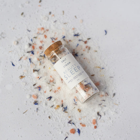 Bottle of 'Beauty Sleep' facial steam on a textured white surface with colourful bath salts around.