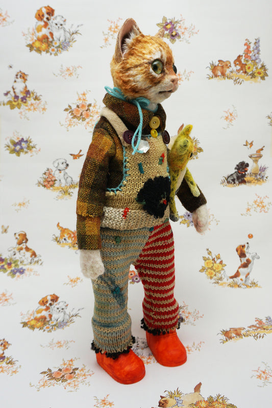 A whimsical cat doll stands upright, wearing a colourful knitted outfit with mismatched patterns and bright orange shoes. It holds a small yellow toy cat. The background features a playful animal-themed pattern.