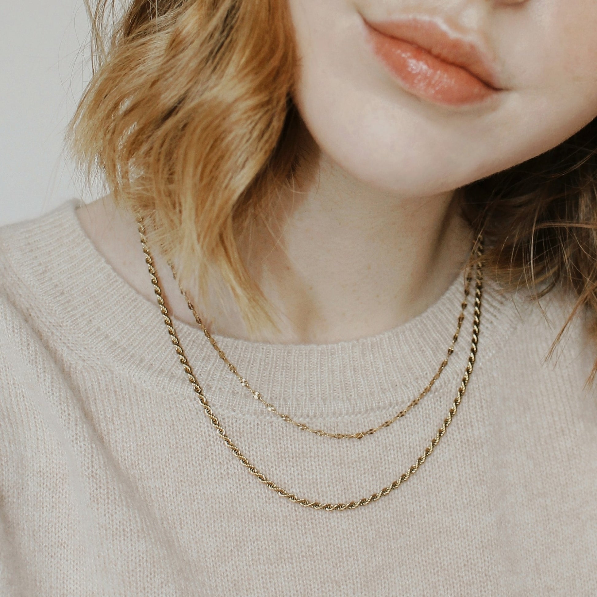 One gold necklace that gives the appearance of two layered gold necklaces on a person wearing a beige jumper.