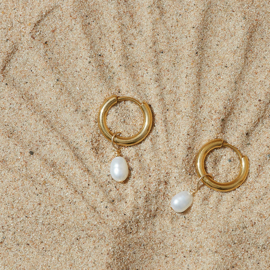 A pair of gold-coloured hoops featuring a single pearl hanging underneath, placed on a sandy surface.
