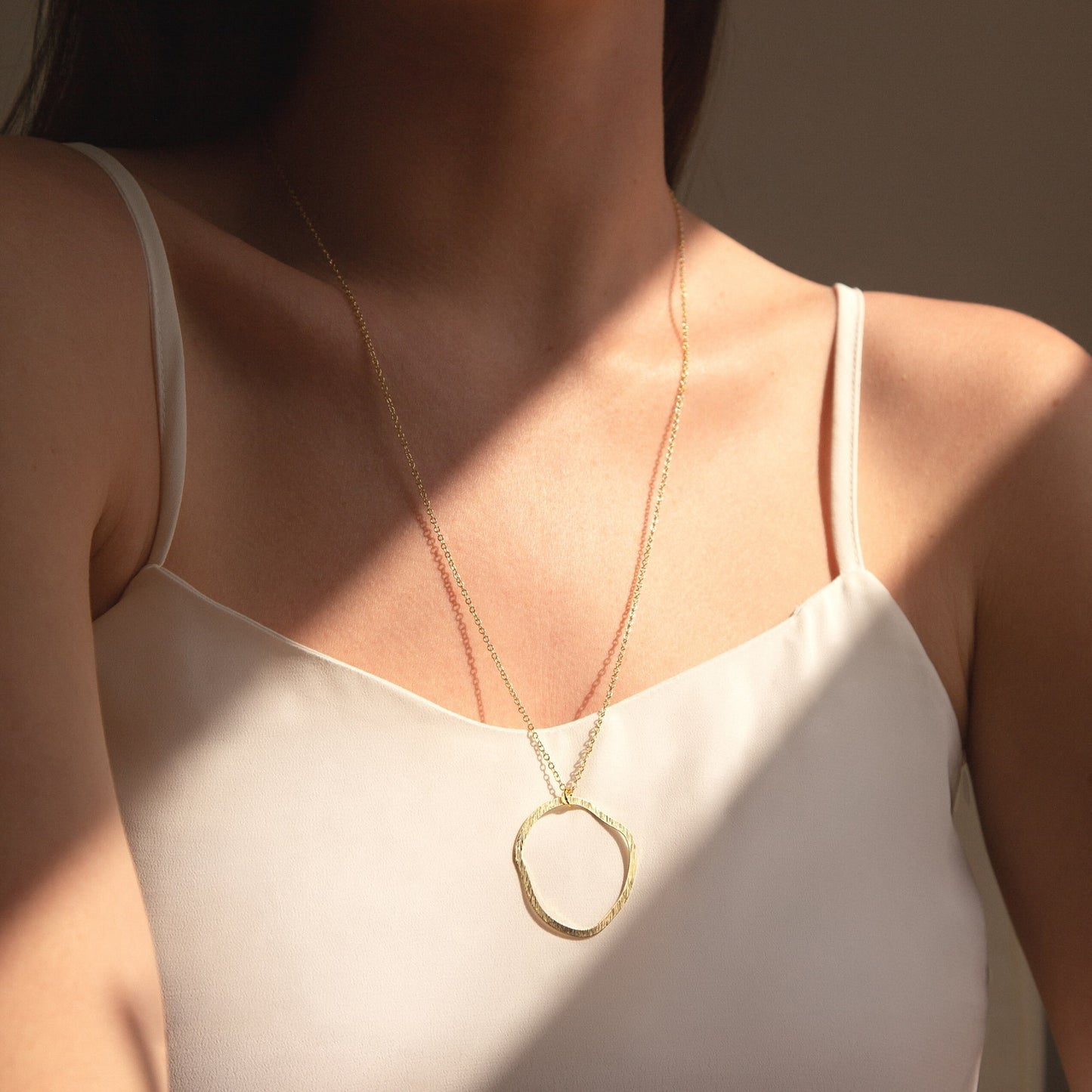 A person wearing a white tank top showcasing a gold-coloured circular line necklace.
