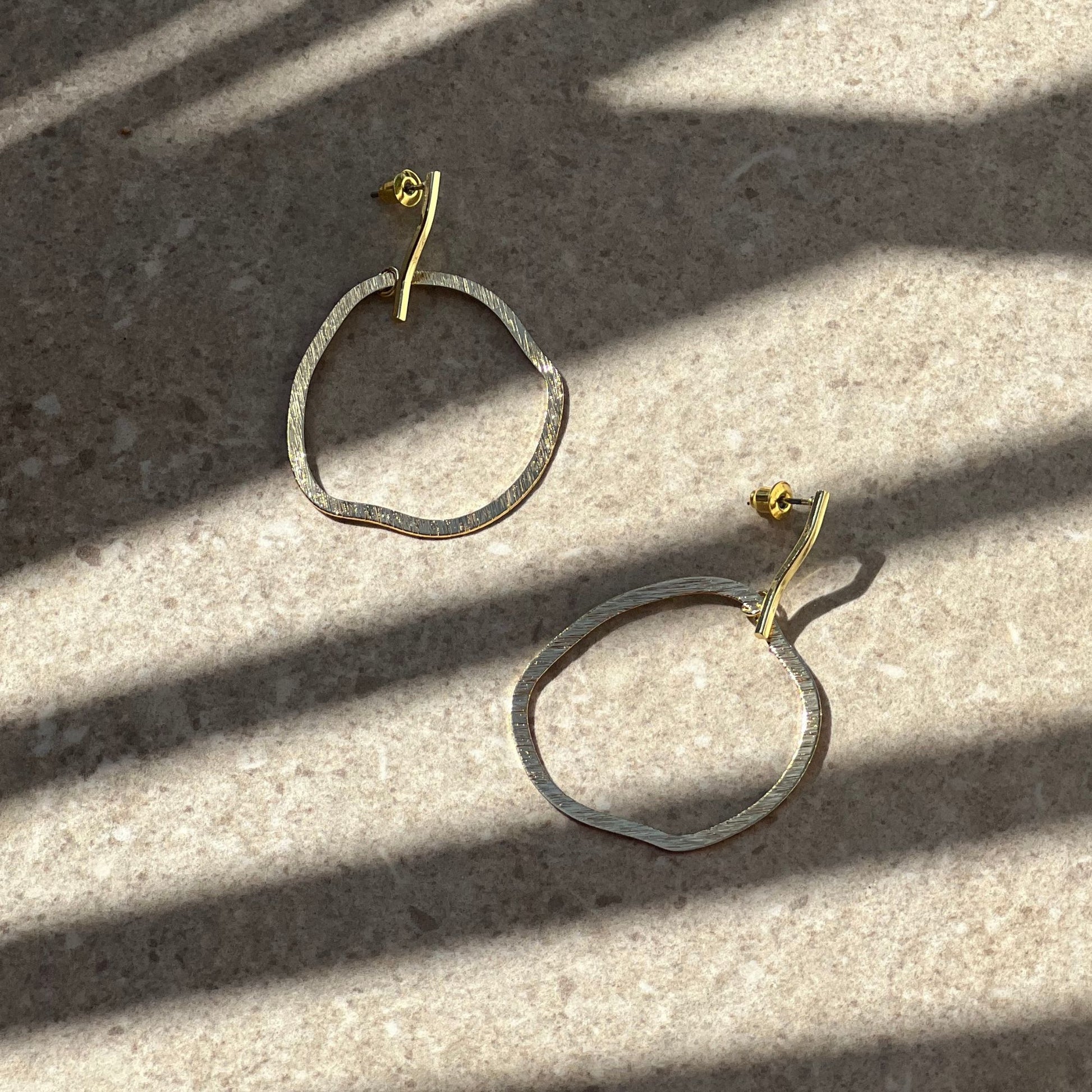 A pair of hoop earrings with a silver and gold color scheme, placed on a surface with natural lighting patterns.